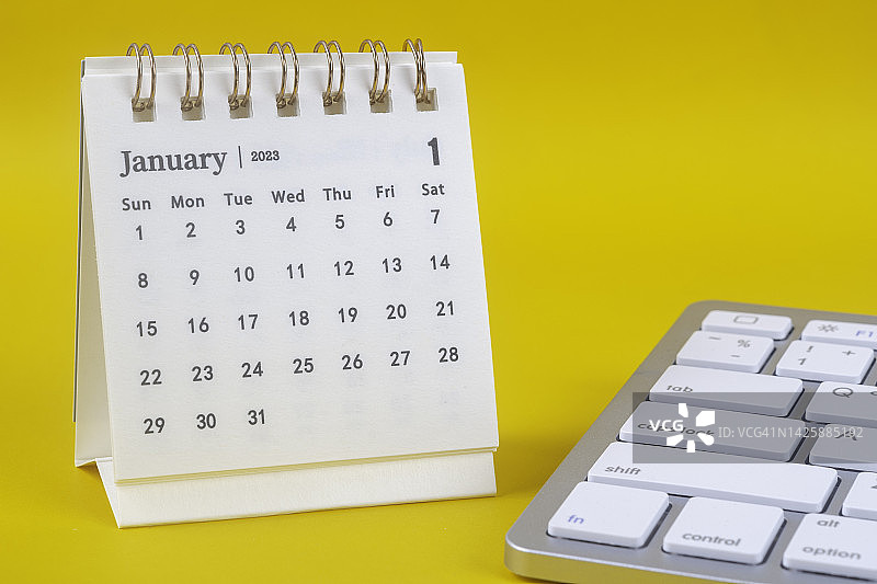 2023日历台:1月是组织者计划和截止日期的月份，白色的电脑键盘和黄色的纸背景。图片素材