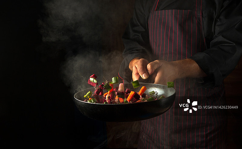 专业厨师的双手将蔬菜块扔进热煎锅中，黑色背景上有蒸汽。酒店烹饪概念图片素材