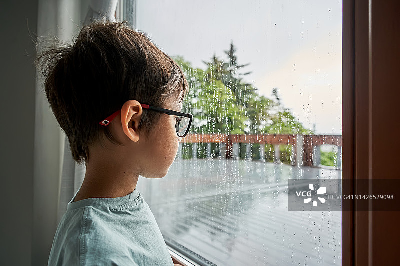 男孩站在窗前看着雨。图片素材