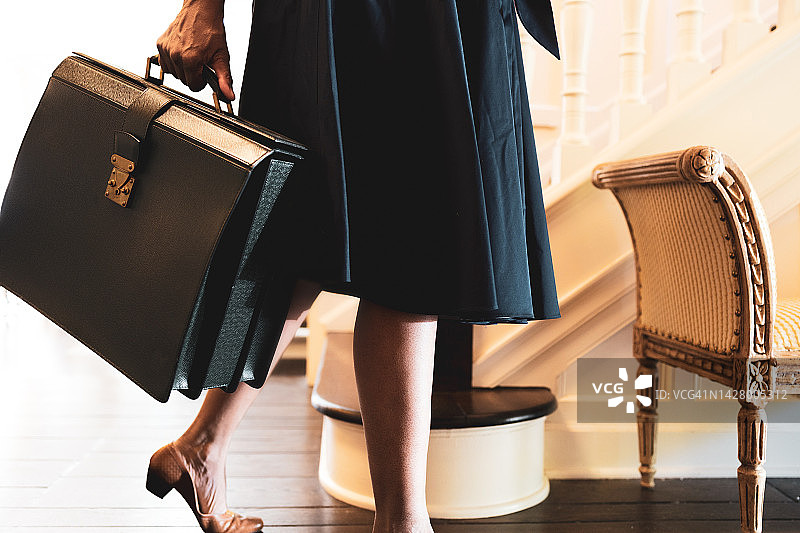 黑人妇女拿着公文包，穿着喇叭裙和高跟鞋站在家里的门厅里图片素材