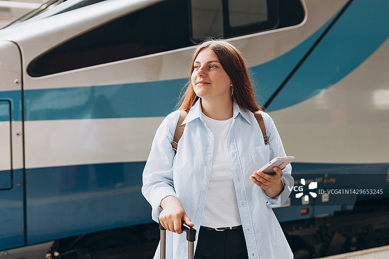 年轻的红发女子背着背包，用智能手机等火车。铁路运输概念，旅行者。提着行李箱的妇女走在火车站站台上。乘火车去度假图片素材