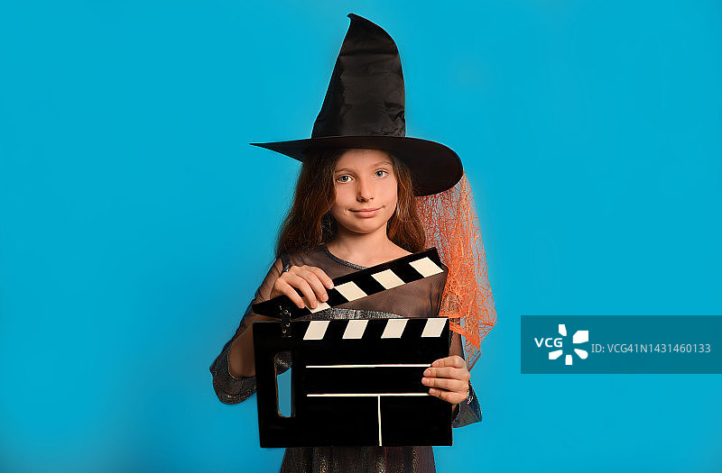 小女巫女孩在万圣节巫师服装手持经典的黑色电影制作隔板，看着相机在蓝色的背景。图片素材