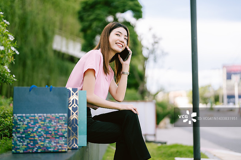 年轻的亚洲妇女拿着购物袋在商场前快乐地坐在电话在早上图片素材