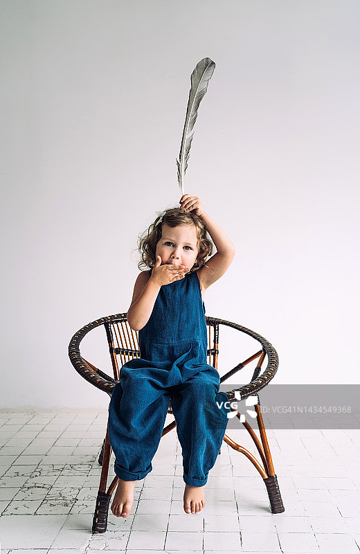 金发卷发的小女孩坐在柳条椅上，头上举着一根羽毛，用手捂着嘴。她假装自己是印第安人。快乐无忧无虑的童年观念图片素材