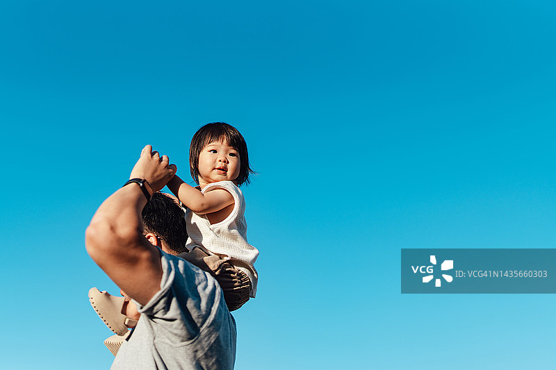 可爱的亚洲幼童坐在父亲的肩膀上，背对着蓝天图片素材