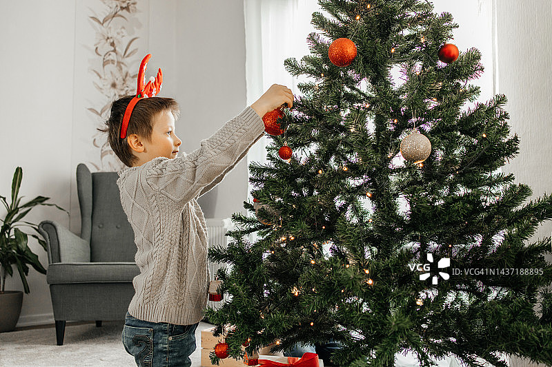 男孩在客厅里装饰圣诞树，在树枝上挂了一个圣诞树玩具图片素材