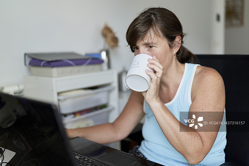 女人穿着便装坐在家里的办公桌前一边喝咖啡一边看电脑图片素材