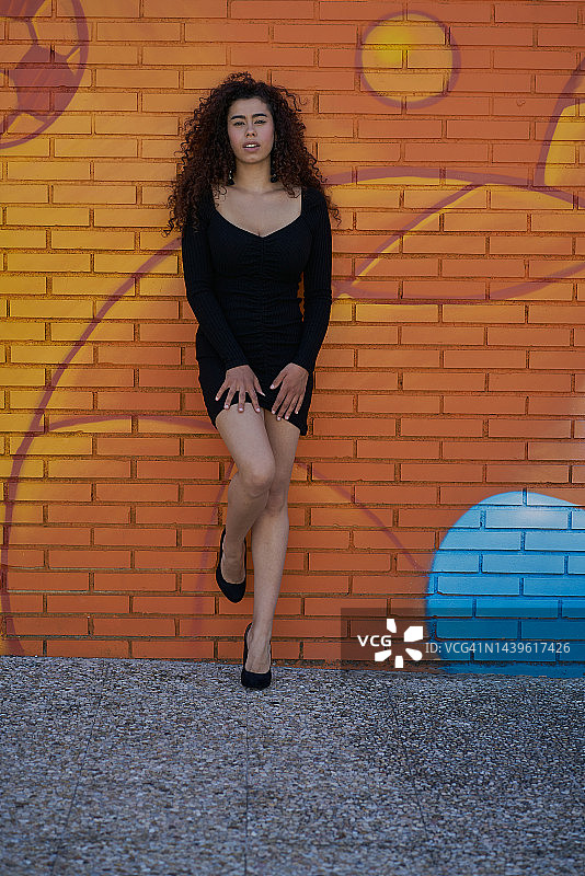 年轻的拉丁女子拿着一双高跟鞋，倚在户外街头的彩色墙壁上摆姿势，墙上画着蓝色的涂鸦。城市的概念。图片素材
