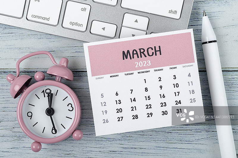 日历桌2023:3月是组织者计划和截止日期的月份，木桌背景上有闹钟和电脑键盘。图片素材