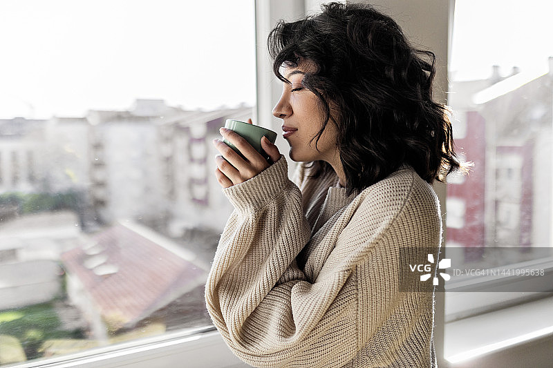 年轻女子一边享受着咖啡，一边从家里的窗户往外看图片素材