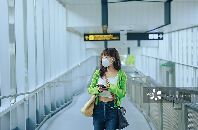 年轻的亚洲女商人戴着口罩拿着手机站在机场离境区。商务旅行的概念。图片素材
