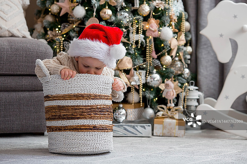 一个漂亮的小男孩戴着圣诞老人的帽子坐在篮子里，在圣诞树的背景下快乐地笑着。新年礼物。庆祝圣诞节的概念。图片素材