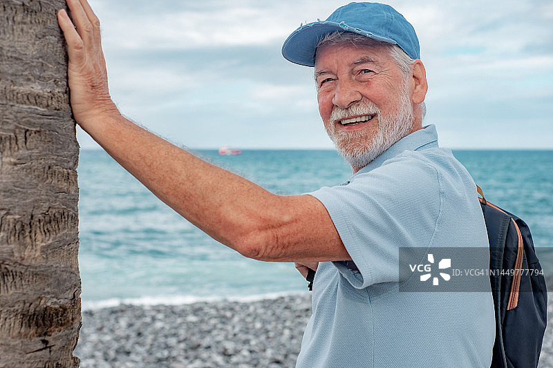 微笑的高级男子站在海滩上，靠近一棵棕榈树，享受空闲时间假期或退休。有吸引力的胡子男子与帽子和白发看着相机。水面上的地平线图片素材