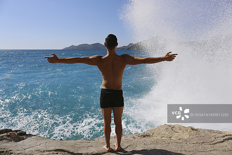 男人张开双臂看着海浪拍打着岩石图片素材