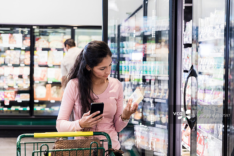拿着手机的女人在食品杂货店冷冻区检查产品标签图片素材