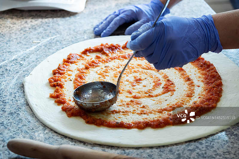 我们在家里、披萨店或咖啡馆做意大利素食披萨。女面包师、糕点师或推销员在面包店用勺子把番茄酱或番茄酱涂在擀好的面团上。食物的概念。一步一步的指导，自己动手。简单的生活。图片素材