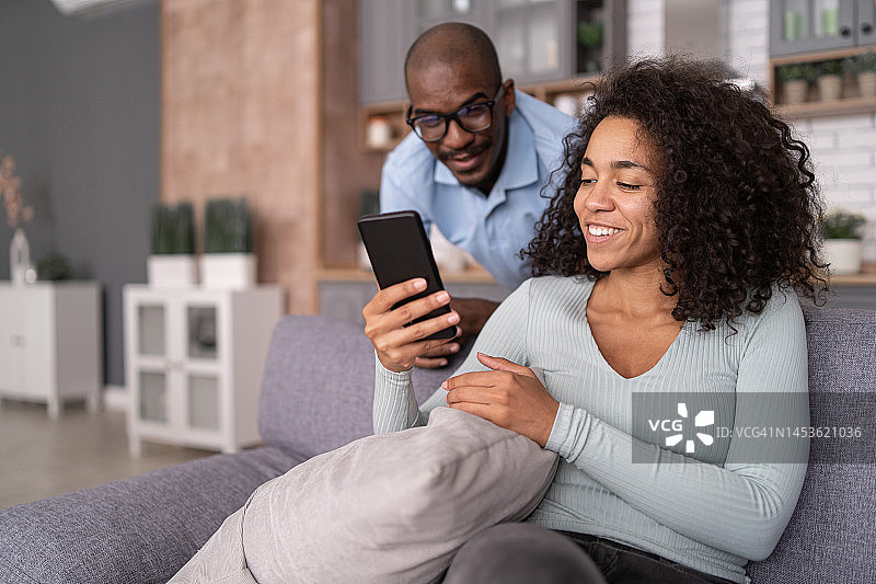 快乐的年轻非裔美国家庭夫妇用手机，笑着看搞笑的在线视频，在线视频通话，坐在家里的沙发上。女人向男人展示她的手机屏幕。图片素材