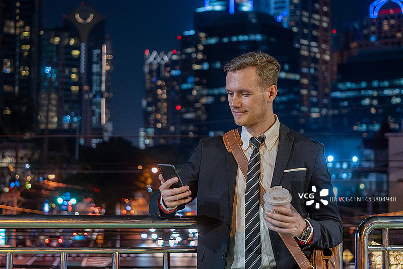 帅哥使用智能手机行走在充满霓虹灯色彩和娱乐的夜晚城市。时髦的年轻人使用手机。图片素材