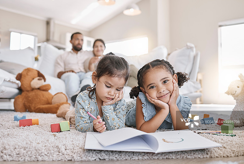 两个小女孩躺在客厅的地板上用彩色铅笔画画，他们的父母在沙发上休息。孩子们，姐妹们，兄弟姐妹在家里的家庭时间里画画图片素材