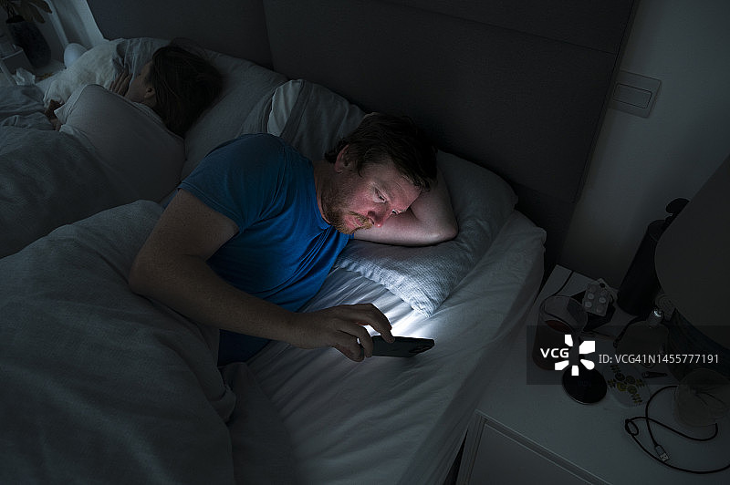 一个成熟的男人醒着躺在床上，而他的伴侣在睡觉。他使用智能手机图片素材