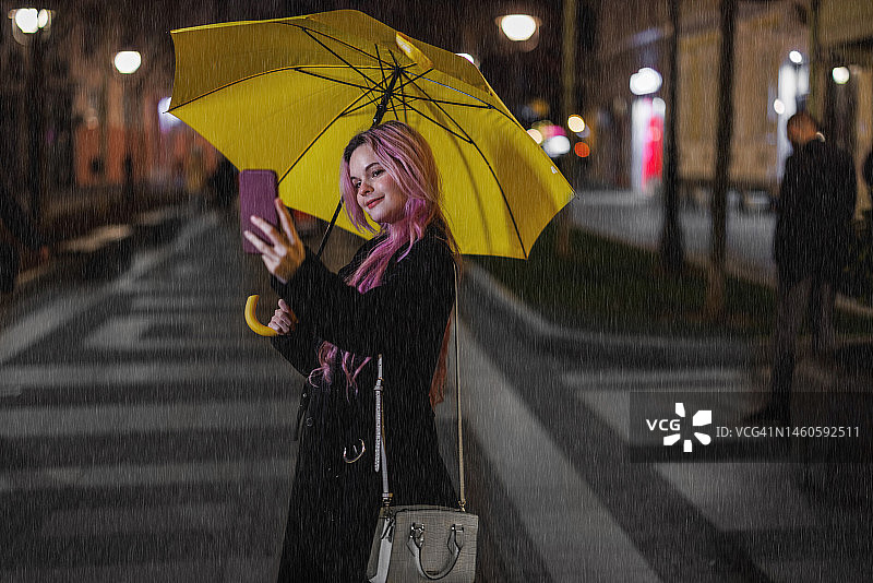雨夜，一名年轻女子打着黄色雨伞走在街上，用手机自拍。图片素材