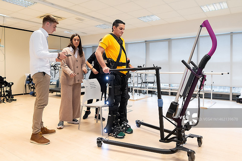 机械外骨骼。现代医院的物理治疗:物理治疗师和科学家用机器人骨架举起残疾人。科学家、工程师和理疗康复医生使用平板电脑提供帮助图片素材