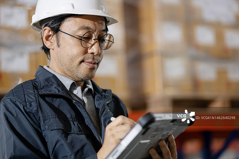 制造业ERP解决方案中的仓库管理系统(WMS)。侧视图的日本仓库经理使用平板电脑监控库存水平和仓库操作的工厂仓库。图片素材