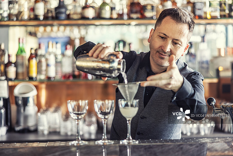 专业的调酒师在吧台准备鸡尾酒，将酒从摇酒器中倒入过滤器中。图片素材