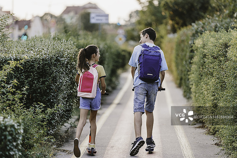 哥哥和姐姐背着背包骑着滑板车在城市里。两个孩子骑自行车去上学。副本的空间。图片素材