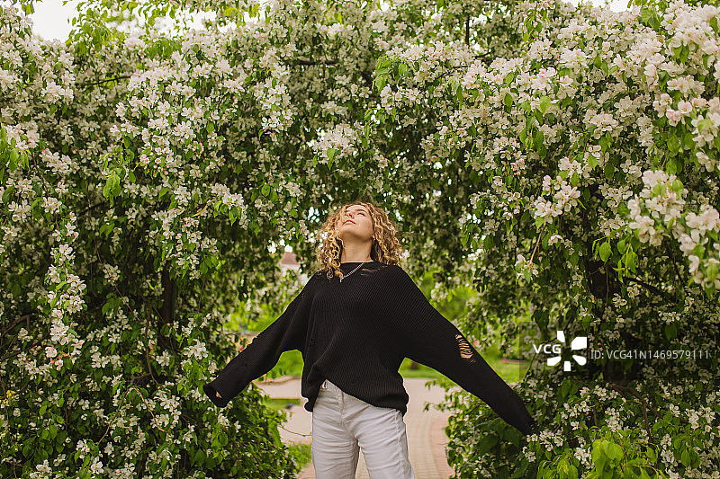 金发卷发的女孩在黑色毛衣的背景上开花的树。花园。白色的花朵图片素材