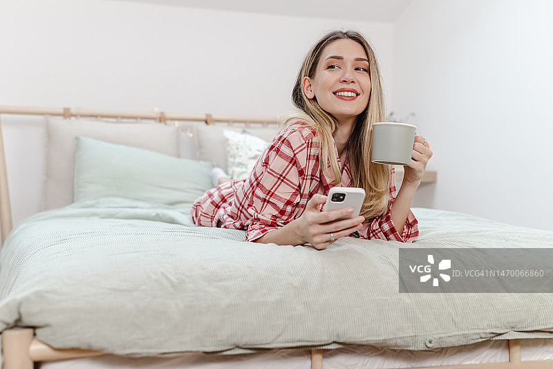 年轻女子早上在床上喝咖啡，用智能手机图片素材