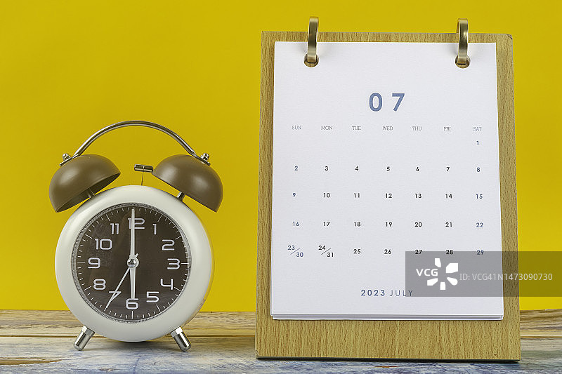 日历台2023:七月是组织者计划和截止日期的月份，在桌子上有一个闹钟，背景是黄色的纸。图片素材