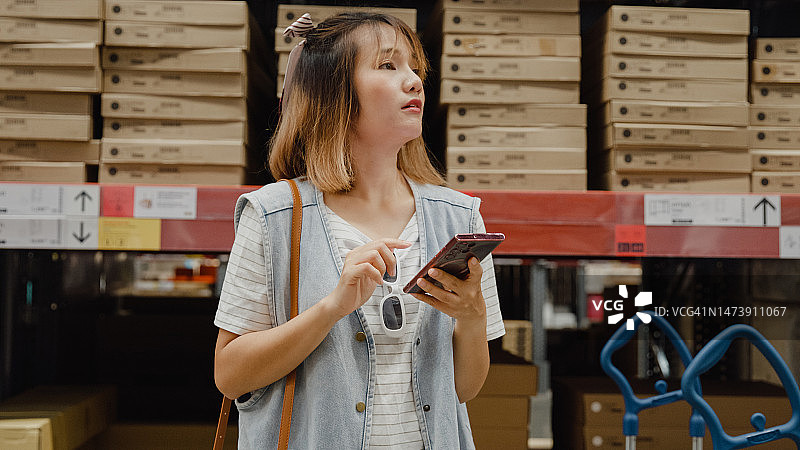 年轻的亚洲女性顾客使用智能手机在零售仓库搜索商品库存。商业仓库购物。图片素材