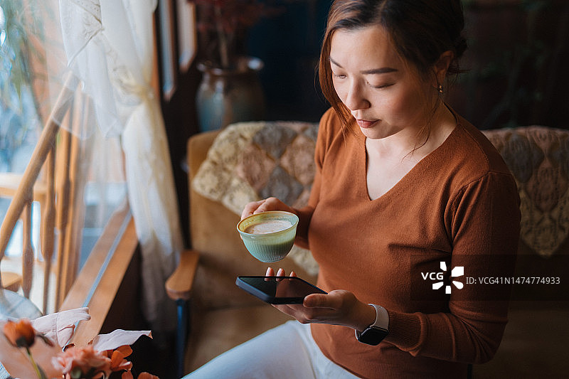 放松的年轻亚洲女性在舒适的家里一边喝着咖啡，一边用智能手机在网上购物。移动购物和电子商务的便利性。非接触式支付概念图片素材