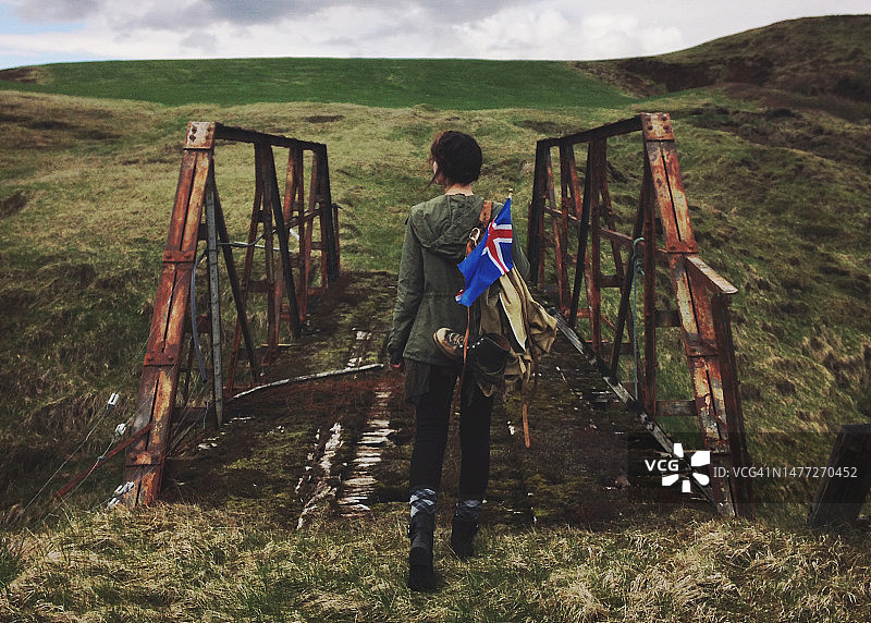 一名女子背着背包、靴子和冰岛国旗，在乡村秋天的风景中穿过一座生锈的桥，冰岛图片素材