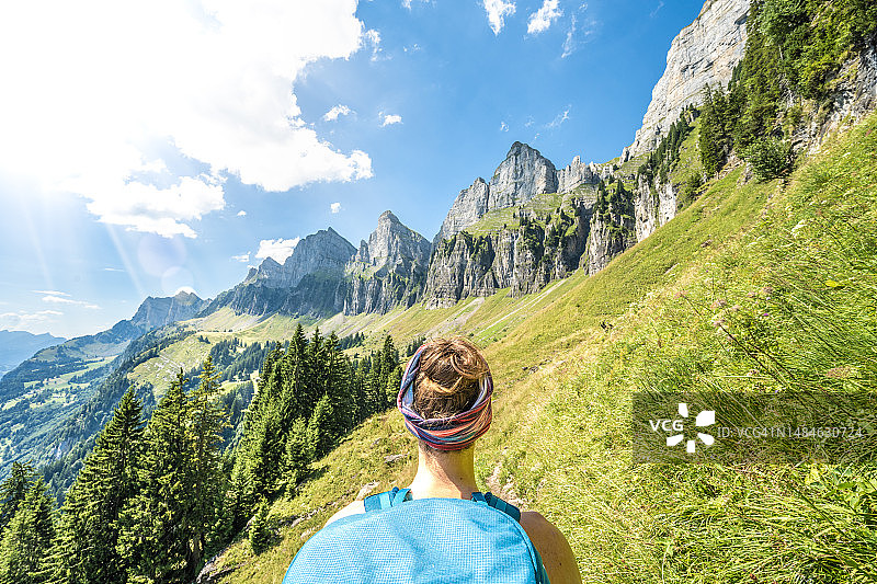 一位女性徒步者在草地和树木之间的徒步小径上的肩部视图，背景是churf<s:1>斯滕山脉。瑞士，瓦伦塞，圣加仑，瑞士，欧洲。图片素材