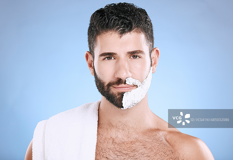 男人的肖像与泡沫在半胡子，毛巾和皮肤植入产品在工作室模拟。剃须膏在脸上，头发和皮肤护理快乐男模美容，隔离在蓝色背景。图片素材