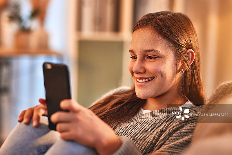 快乐的少女周末在家放松地躺在沙发上玩手机、社交媒体和在线交流。年轻的女性面带微笑，在客厅用手机聊天或者上网玩游戏图片素材