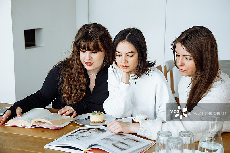 三个女大学生坐在桌子旁，桌上放着蛋糕，打开书和杂志，看着图片图片素材