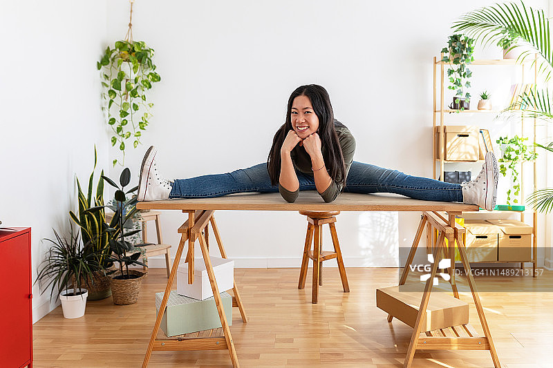 快乐的年轻亚洲女子在家庭办公室的木桌上做瑜伽姿势时对着镜头微笑。图片素材
