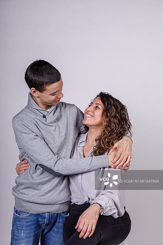 欢快的十几岁的男孩和他的母亲站在白色的背景下，拥抱和亲密图片素材