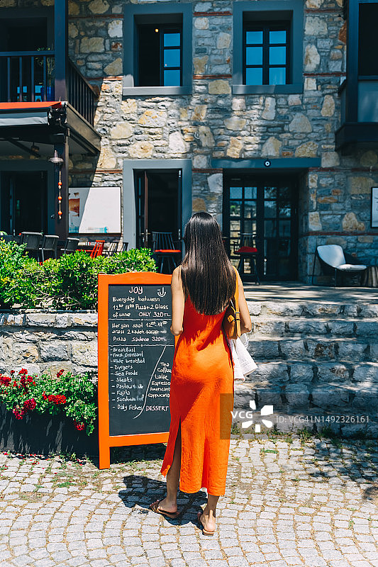 美丽苗条的亚洲女人穿着红裙子在街边咖啡馆读菜单(后视图)。图片素材