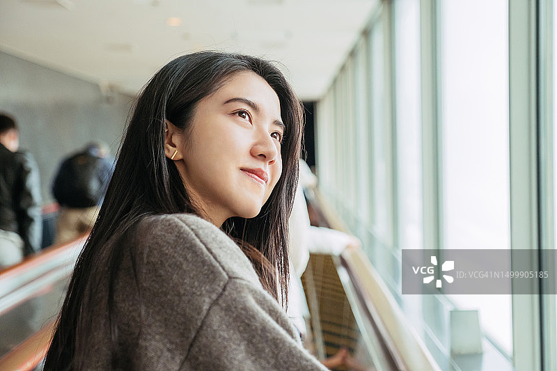 日本大阪，一名年轻美丽的亚洲女子坐在购物中心的自动扶梯上，面带微笑地望向别处。阳光透过窗户照进来。旅游理念，未来与希望。图片素材