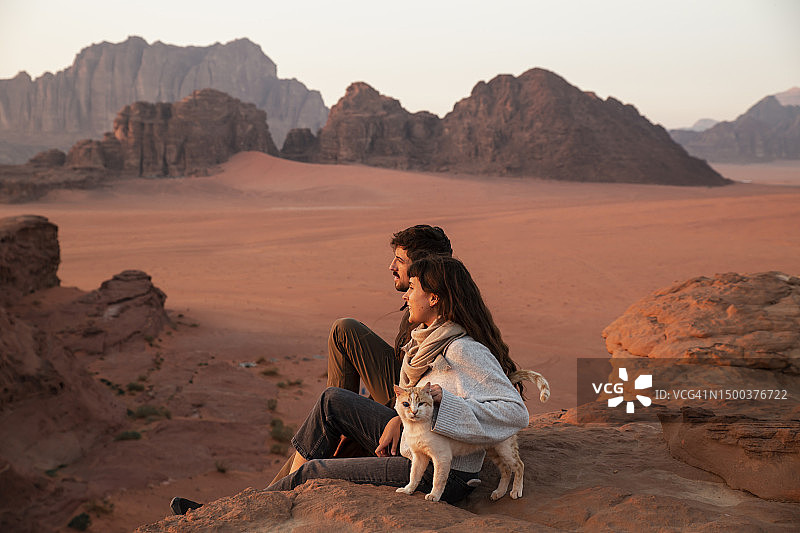 日落时分，一对年轻夫妇和猫坐在沙漠岩石上图片素材