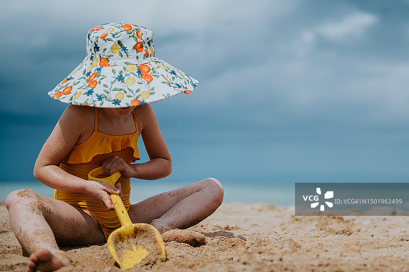 一个小女孩坐在沙滩上，用一把黄色的塑料铲子在沙子里挖图片素材