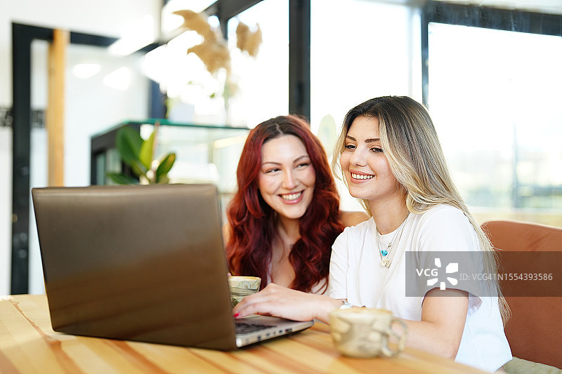 两个年轻女子坐在咖啡馆里用笔记本电脑看视频，喝着咖啡，玩得很开心图片素材