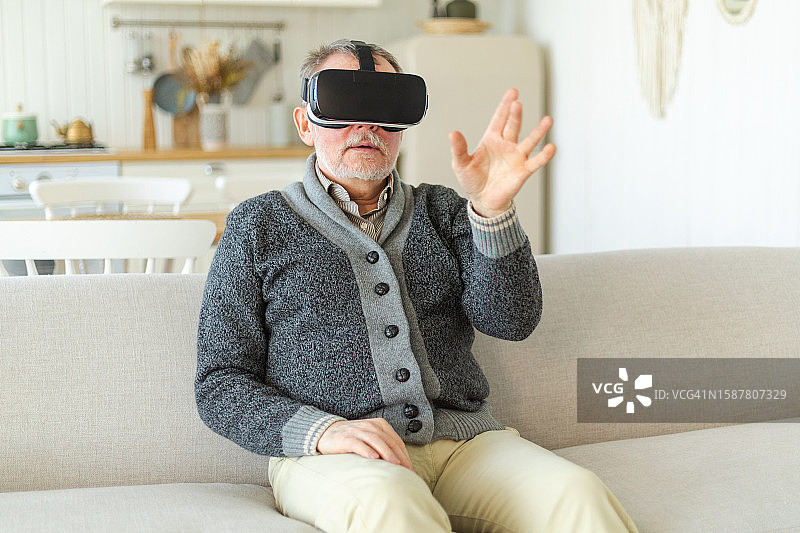兴奋的成熟老年男子在家中戴着使用虚拟现实的虚拟现实头盔。爷爷在虚拟现实头盔上体验VR时触摸空气。模拟高科技视频游戏。图片素材