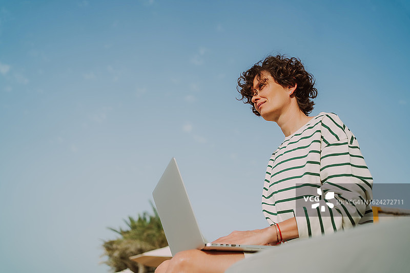 平静的黑发短发的女人把笔记本电脑放在膝盖上，对着蓝天和棕榈树休息。低角度视图图片素材