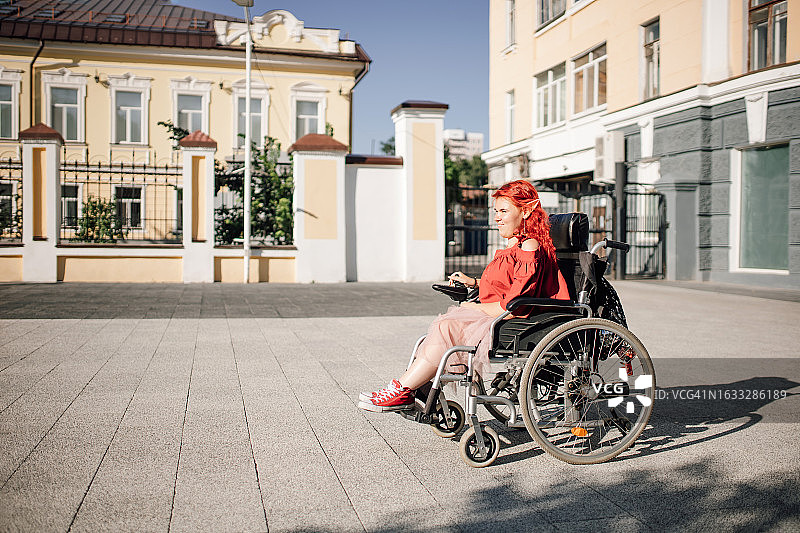 可爱的红头发女人的侧视图享受阳光明媚的日子，坐在轮椅上沿着城市街道骑行图片素材