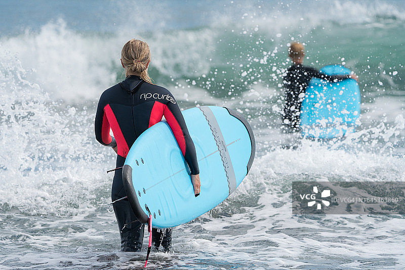 后视图:年轻女子冲浪者穿着潜水服，手里拿着冲浪板，走进太平洋的巨浪中图片素材